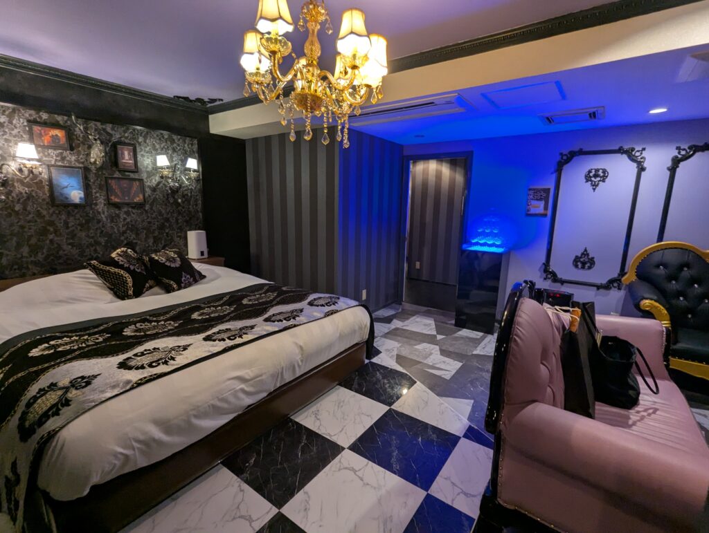 HOTEL LUNA町田店の501号室。手前にゴシック調の豪華なキングサイズベッドがあり、白と黒の市松模様(チェッカーフラッグ柄)の床が広がる。天井にはシャンデリアがきらめき、青い間接照明が部屋の奥を照らしている。