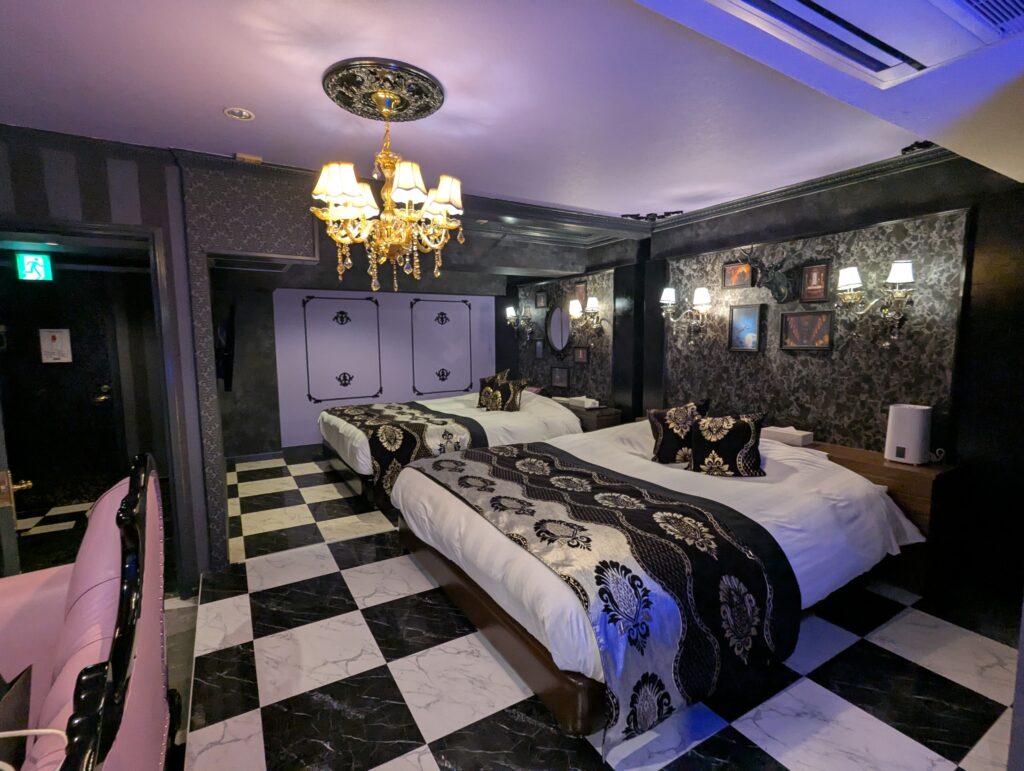HOTEL LUNA町田店 501号室に完備された2つの広々としたダブルベッド。黒とゴールドのゴシック調のベッドライナーとクッションが交互に配置され、ダマスク柄の壁紙とシャンデリアがラグジュアリーな雰囲気を演出している。