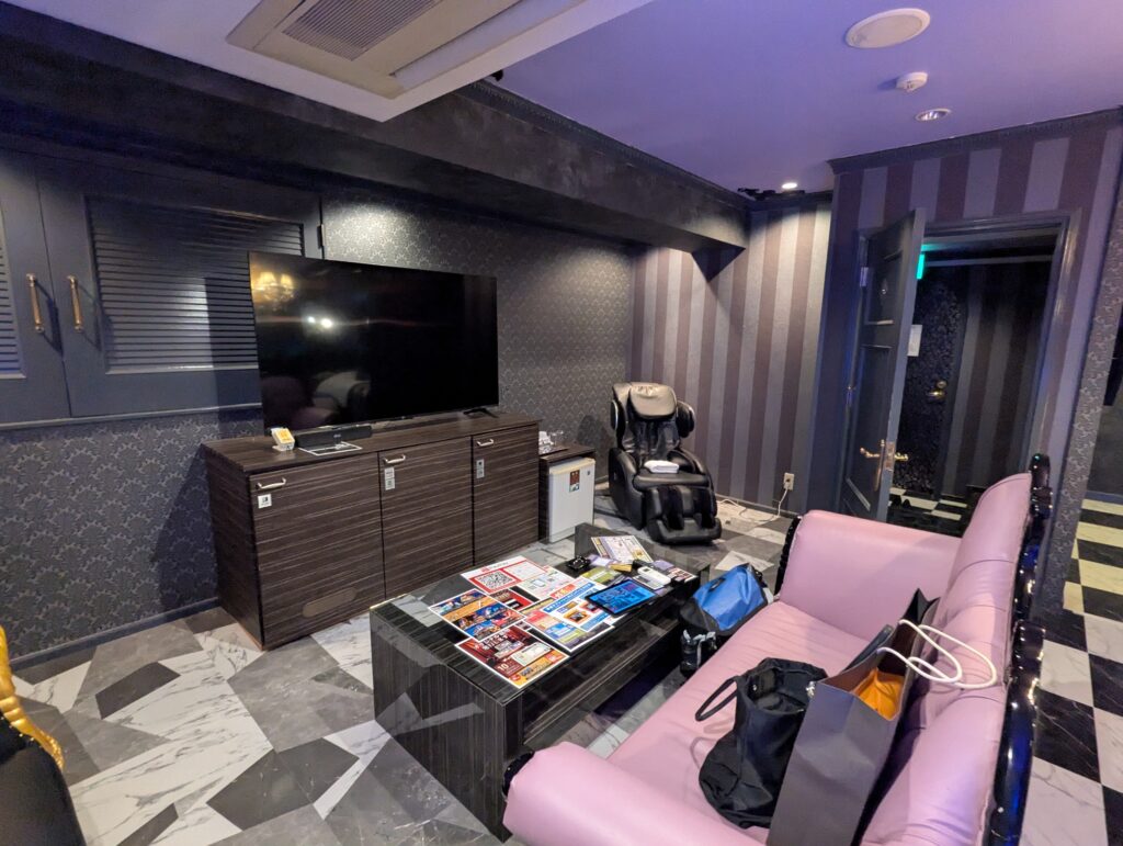 HOTEL LUNA町田店 501号室の大型テレビとリビングスペース。手前にはピンク色のクラシックなソファ、奥には高級感のある黒いマッサージチェアや冷蔵庫が設置されている。壁紙はグレーのストライプとダマスク柄。