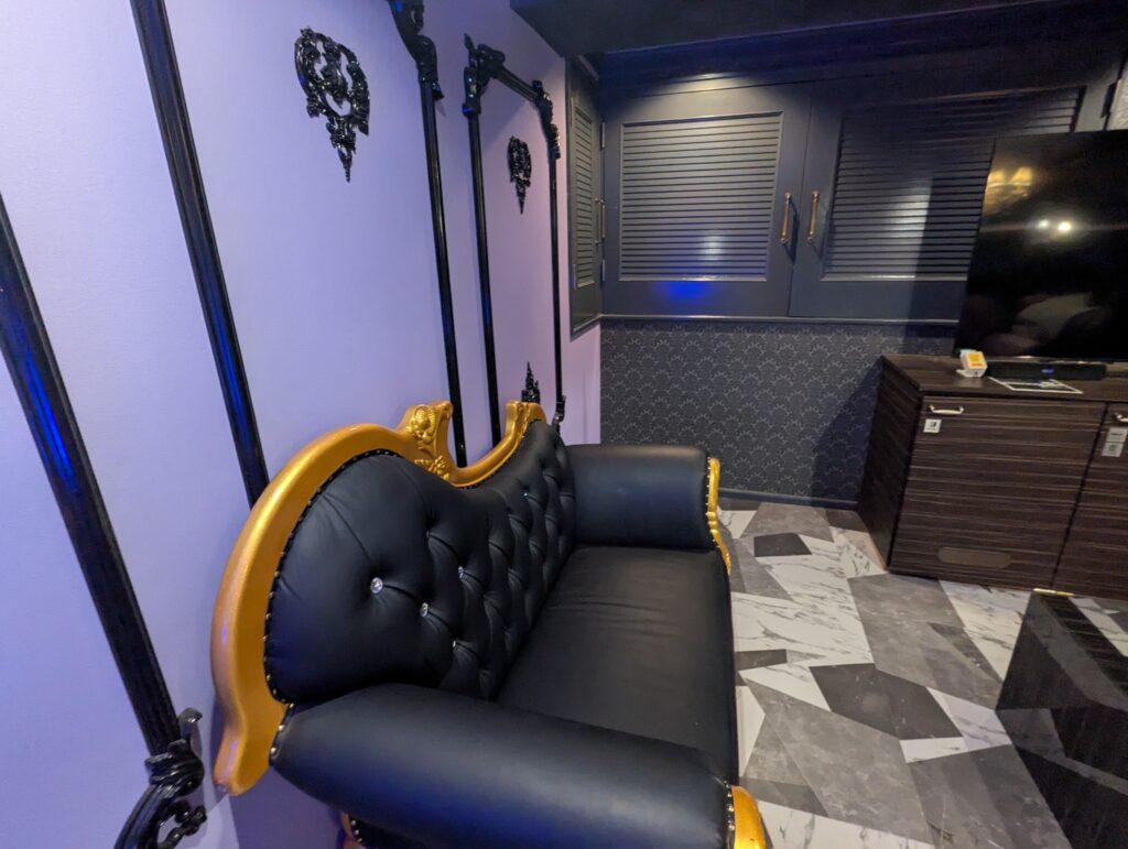 HOTEL LUNA町田店 501号室の壁際に置かれたゴシック調のレザーソファ。ゴールドのフレームに黒いボタン留めの背もたれが特徴的で、薄紫色の壁面には黒いクラシカルな額縁風の装飾が施されている。