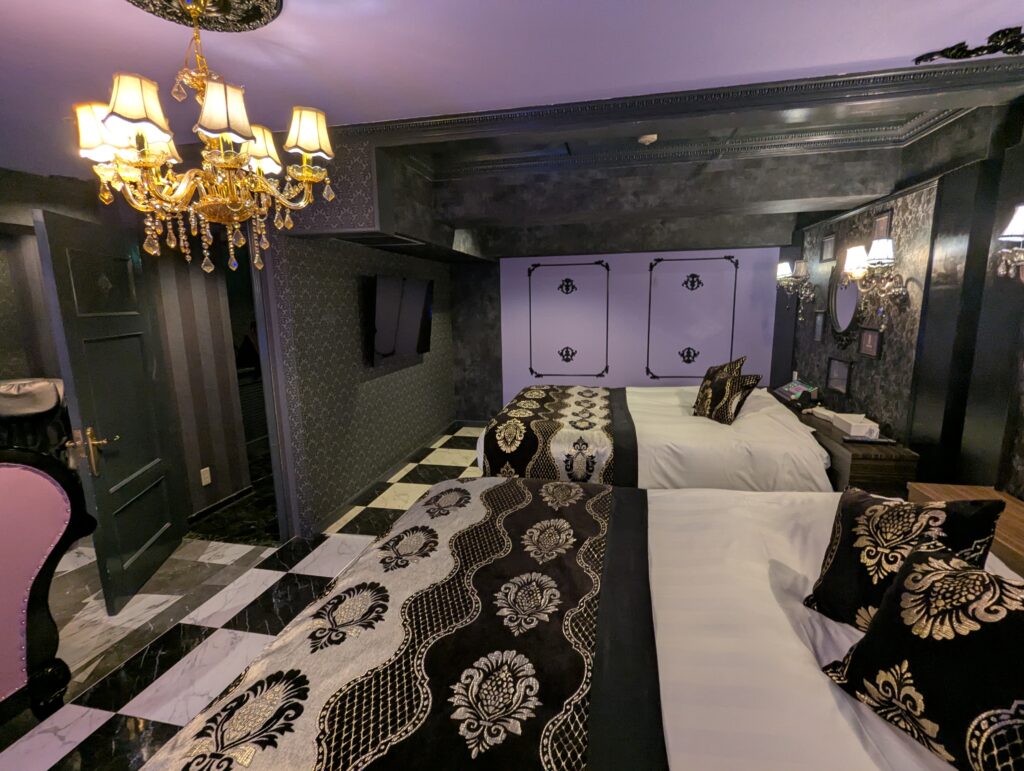 手前から奥に向かって2台のベッドが並ぶ、HOTEL LUNA町田店 501号室のベッドルーム。シャンデリアの温かい光と、部屋全体のダークトーンなゴシックインテリアが絶妙にマッチしている。