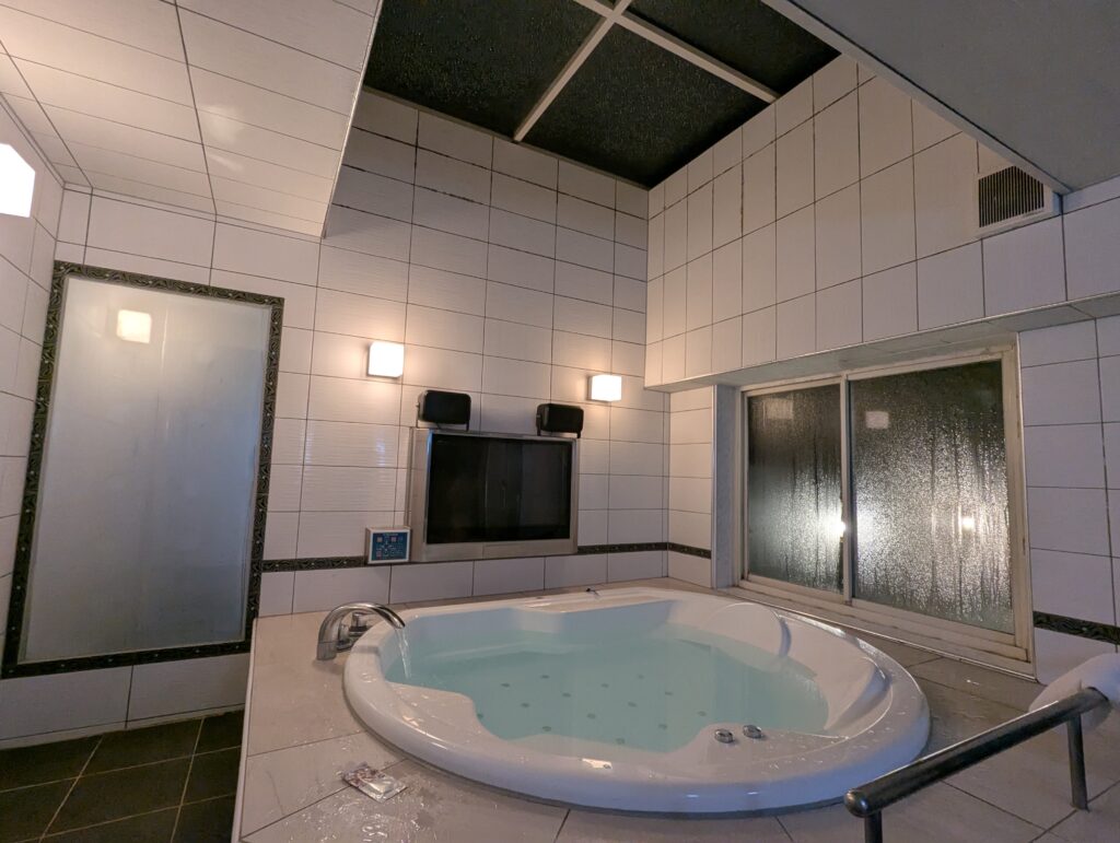 HOTEL LUNA町田店 501号室の広々とした円形ジャグジー付き内風呂(夜の様子)。白いタイル張りの清潔感ある浴室で、浴槽の正面には大型の浴室テレビが設置されている。天井が高く開放感がある。