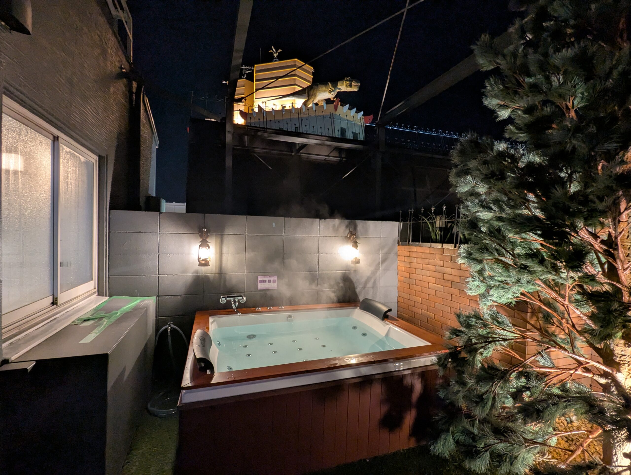 HOTEL LUNA町田店 501号室の木枠の露天風呂(夜)。湯気が立ち上るジャグジーの向こうには、ライトアップされた隣の建物の恐竜オブジェが見えるユニークなロケーション。手前には庭木が植えられている。
