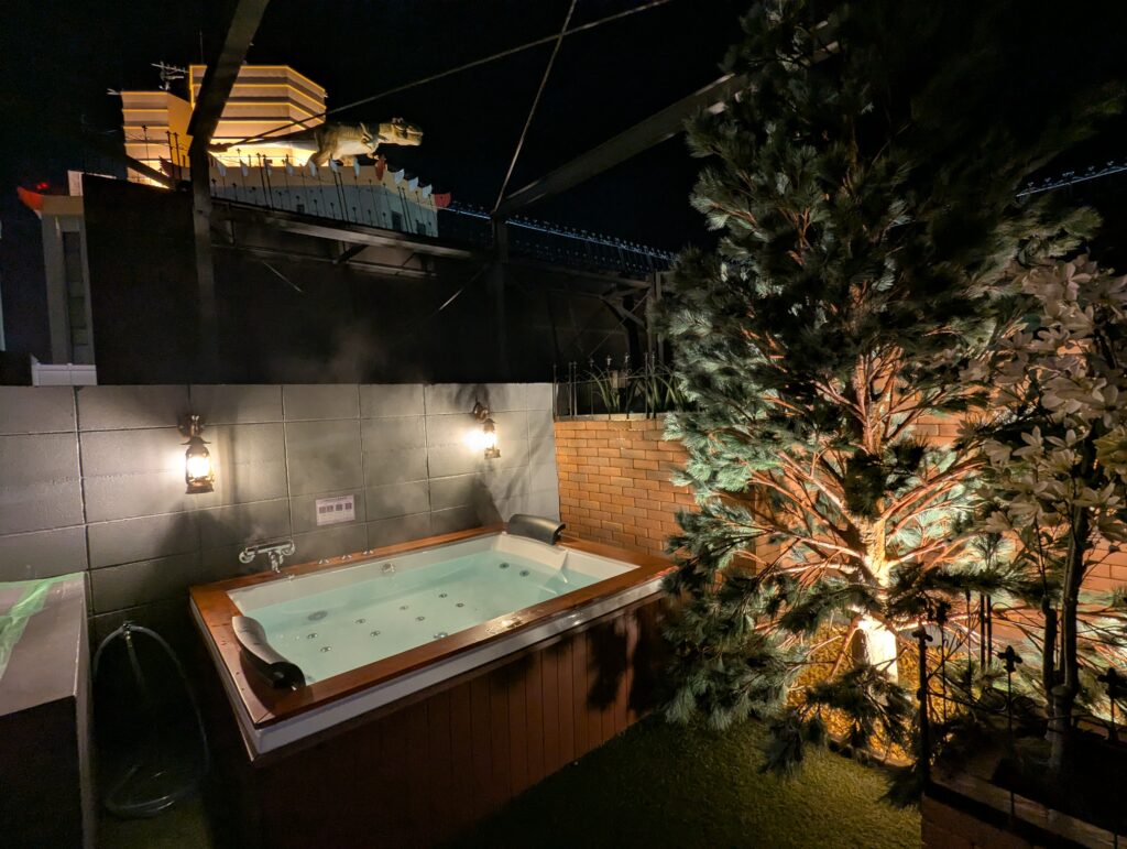夜風を感じられるHOTEL LUNA町田店 501号室の高級感あふれる露天風呂。ウッド調の浴槽パネルと人工芝、レンガ調の壁にアンティーク風のランプが灯り、非日常的な空間を演出している。