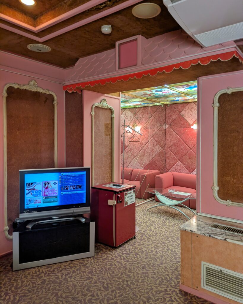 ホテルロンシャン 313号室のテレビ・リビングスペース。ブラウンの壁に囲まれた空間に液晶テレビと赤い小型冷蔵庫が置かれている。奥の部屋にはピンク色のソファとガラスのローテーブルがあり、天井にはキャラクターが描かれたカラフルなライト付きパネルが見える。