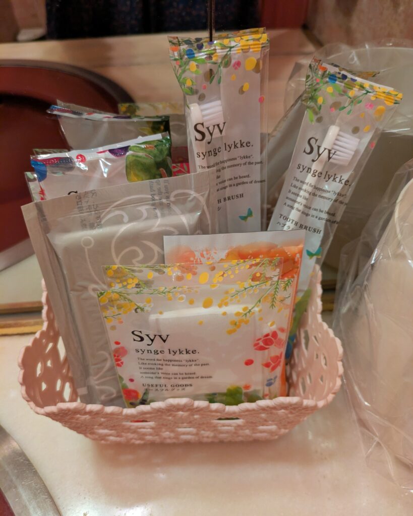 洗面台に用意されたアメニティグッズ。ピンク色のプラスチック製カゴの中に、「Syv synge lykke.」と書かれた北欧風デザインのパッケージに入った歯ブラシやコットンなどのアメニティが綺麗にまとめられている。
