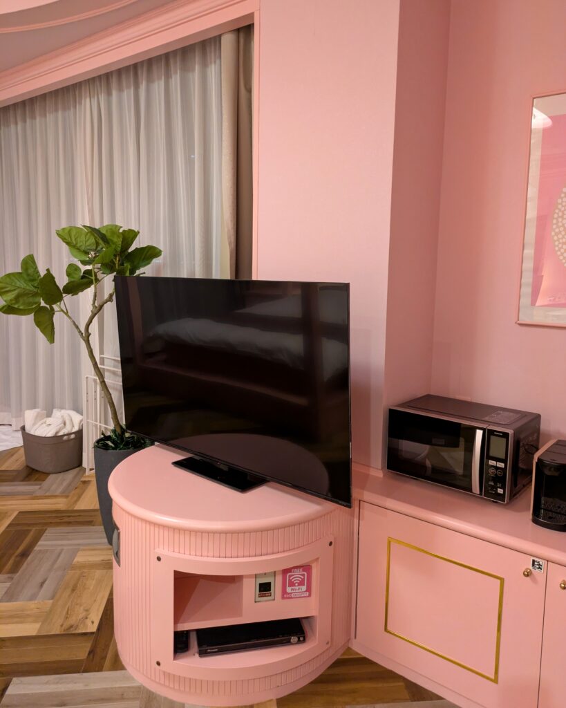 ピンク色の丸いテレビ台に設置された大型液晶テレビ。右隣のカウンターには電子レンジが置かれており、部屋の随所に観葉植物やアメニティが配されている。