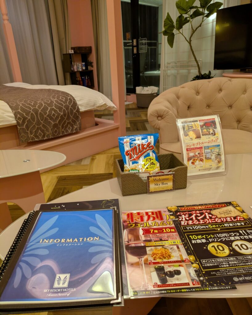 客室のガラステーブルの上に置かれたホテルのインフォメーション冊子やウェルカムサービスの案内POP。カゴの中には無料サービスと思われるマイクポップコーンの袋が入っている。