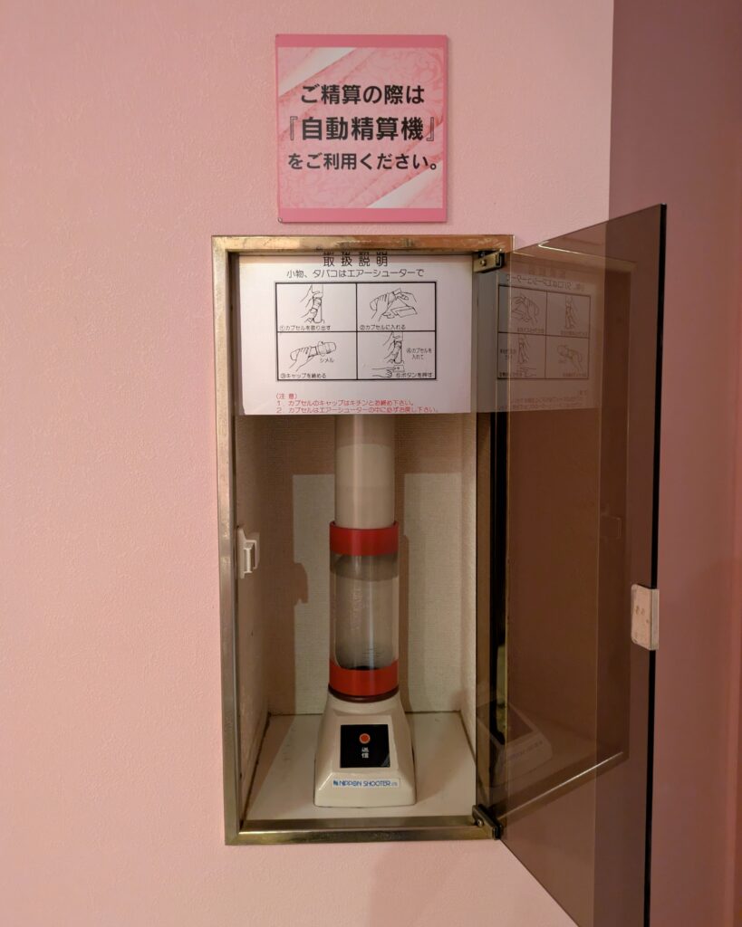 昭和レトロな雰囲気を残す、壁に埋め込まれたカプセル式のエアーシューター(自動精算用機器)。上部には『ご精算の際は自動精算機をご利用ください』とのピンク色の案内表示がある。