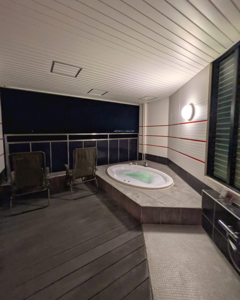 スカイクラブ4 TowerSweet 803号室の露天風呂テラス。夜、青白い光にライトアップされたジャグジー浴槽と、その隣に並ぶ2脚のアウトドアチェア。手前はウッドデッキで、奥には夜の海と遠くの街明かりが見える。オーシャンビューの開放的な空間。